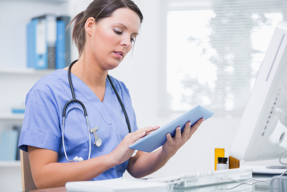 Side View Of Young Female Surgeon Using Digital Tablet In Front Of Computer At Desk In Clinic